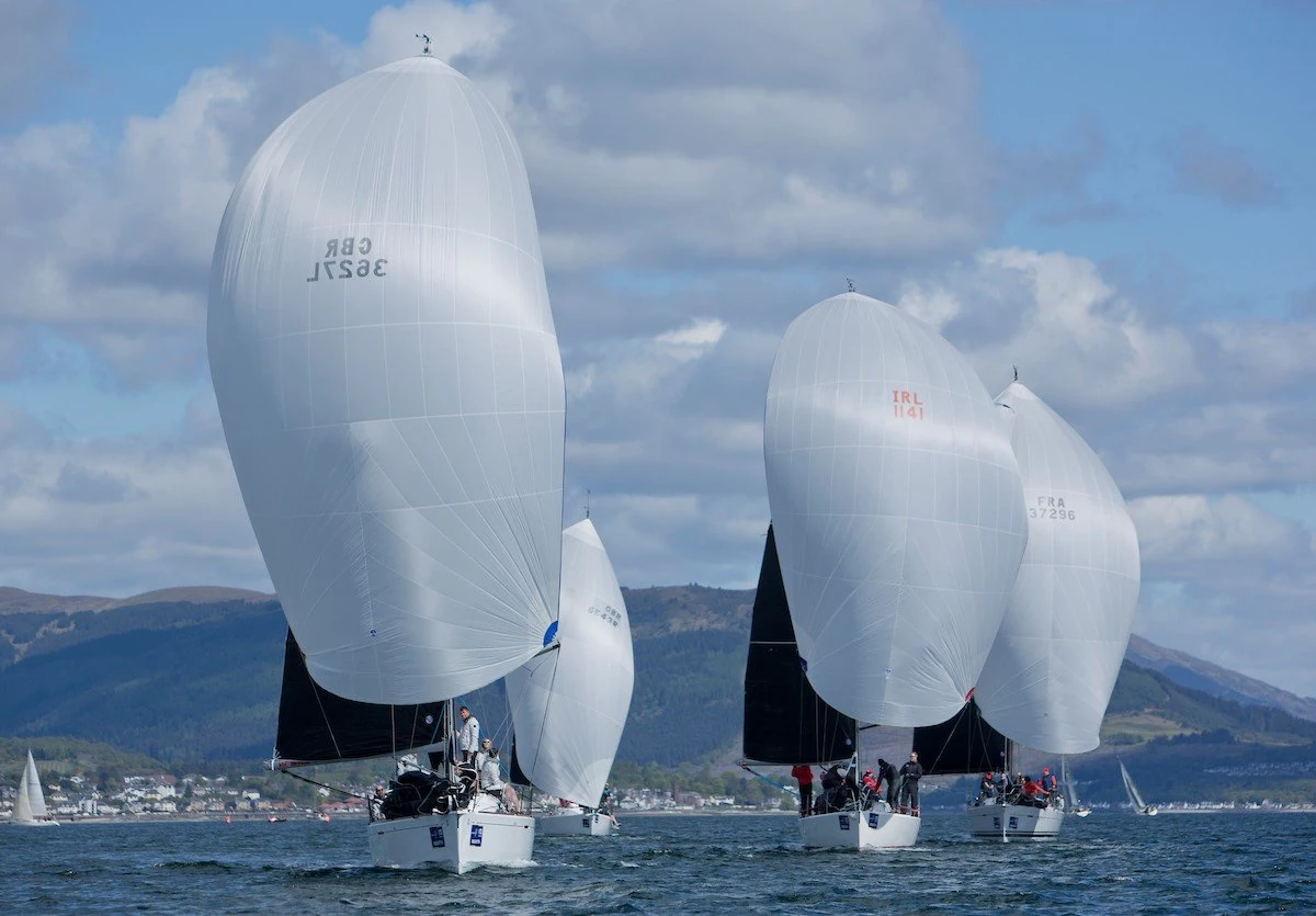 Yachts on the water with pale sails filled with wind, green landmass visible in the background