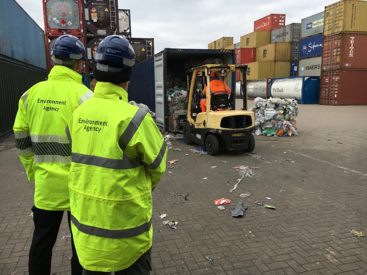 Environment Agency staff in signature yellow jackets stand near an open container or trailer into which a vehicle is positioning itself to pick up material