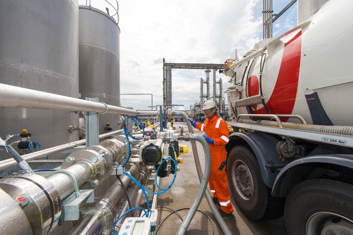 Large steel cylindrical storage tanks at left, and a tanker vehicle with accompanying fuel lines connected to nearby pipes, and an orange-suited worker standing nearby