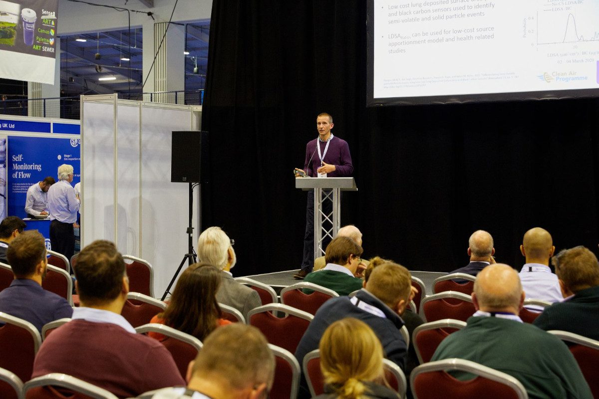 In the background, a darkly-dressed person stands at a lectern in front of an overhead projector screen, in a trade show setting, with audience members visible in front