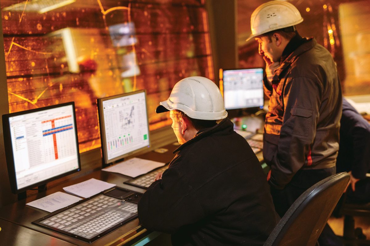 People sitting in a control room looking at computers. Sparks are visible through a nearby window, suggesting they are inside a steelmaking facility