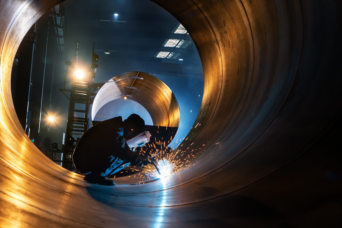 Welder working within a tunnel-like structure made of steel plates with dark industrial interior visible behind the worker, at the opening of this tube-like structure