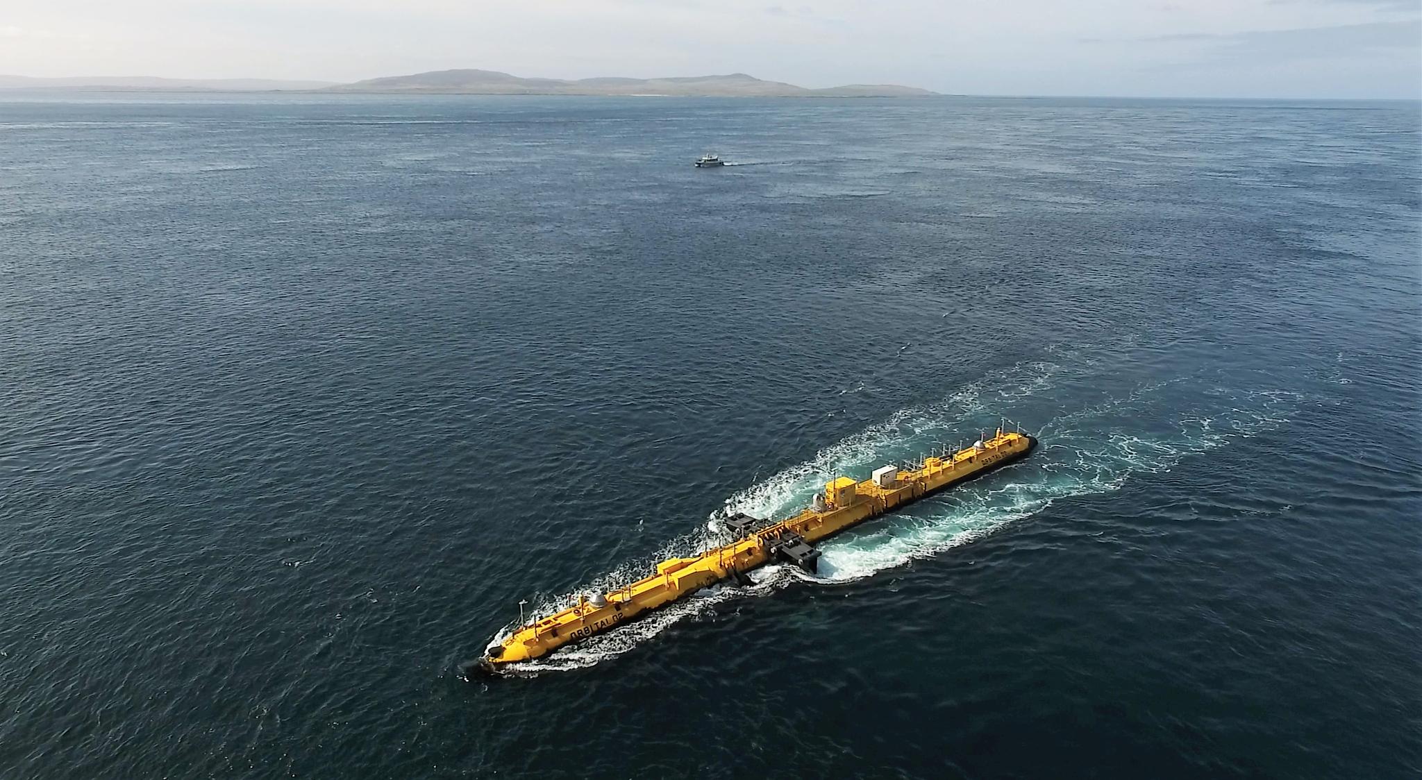 Aerial view of a long, thin, yellow structure sitting in an expanse of sea, with islands in the distance