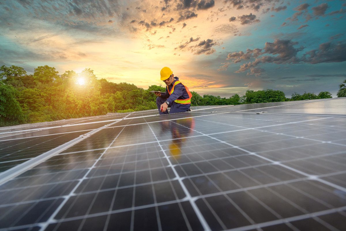 Sloping rooftop installation of solar panels with engineer in the background and dramatic sun visible through trees near the horizon