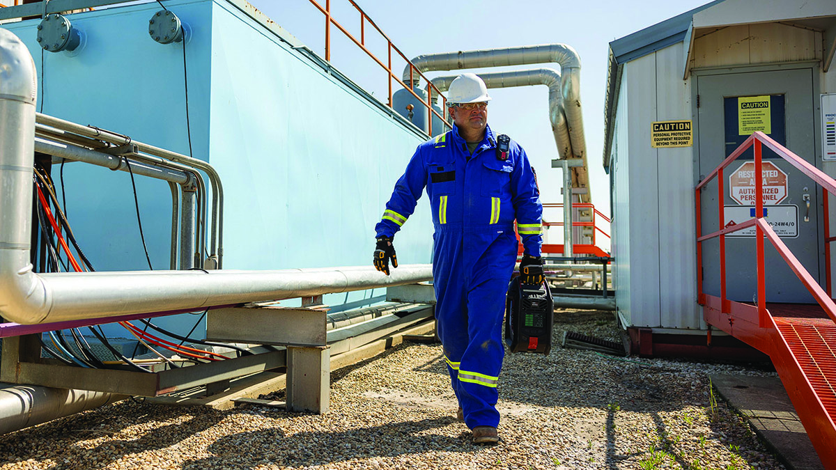 Man in boiler suit and hard hat in safety-critical industrial setting, carrying a dark coloured box, the EXO 8 portable monitor