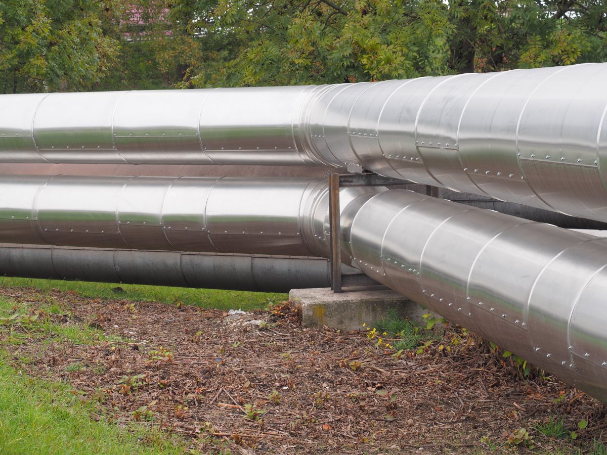 A bend in a system of silver-coloured pipes - district heating pipes - seemingly on a lawn or similar environment