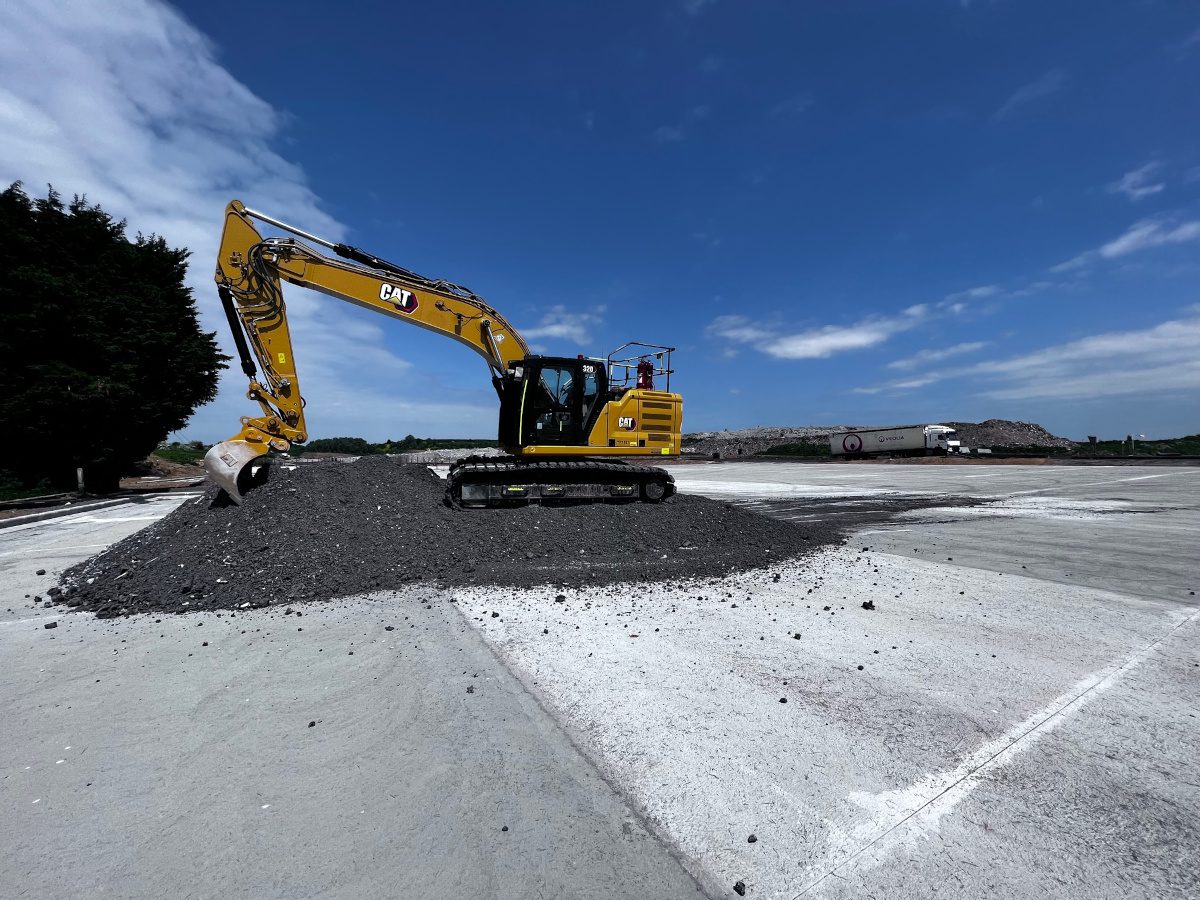 Excavator atop a pile of grey ash within a large flat concrete area with trees and blue sky backdrop