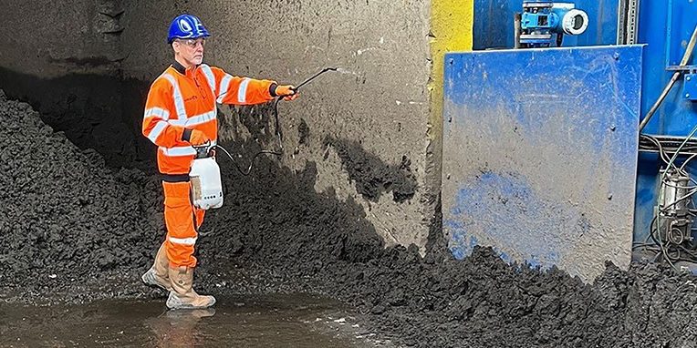 Technician in orange boiler suit spraying an area of a yard, presumably used for storing soil or similar waste material