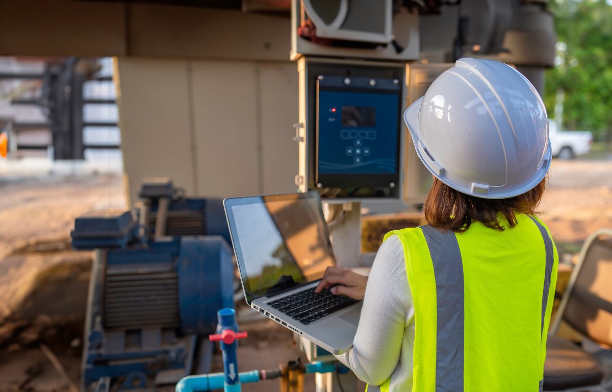 A female wastewater engineer looks at a small display at eye level while holding and typing on a laptop computer