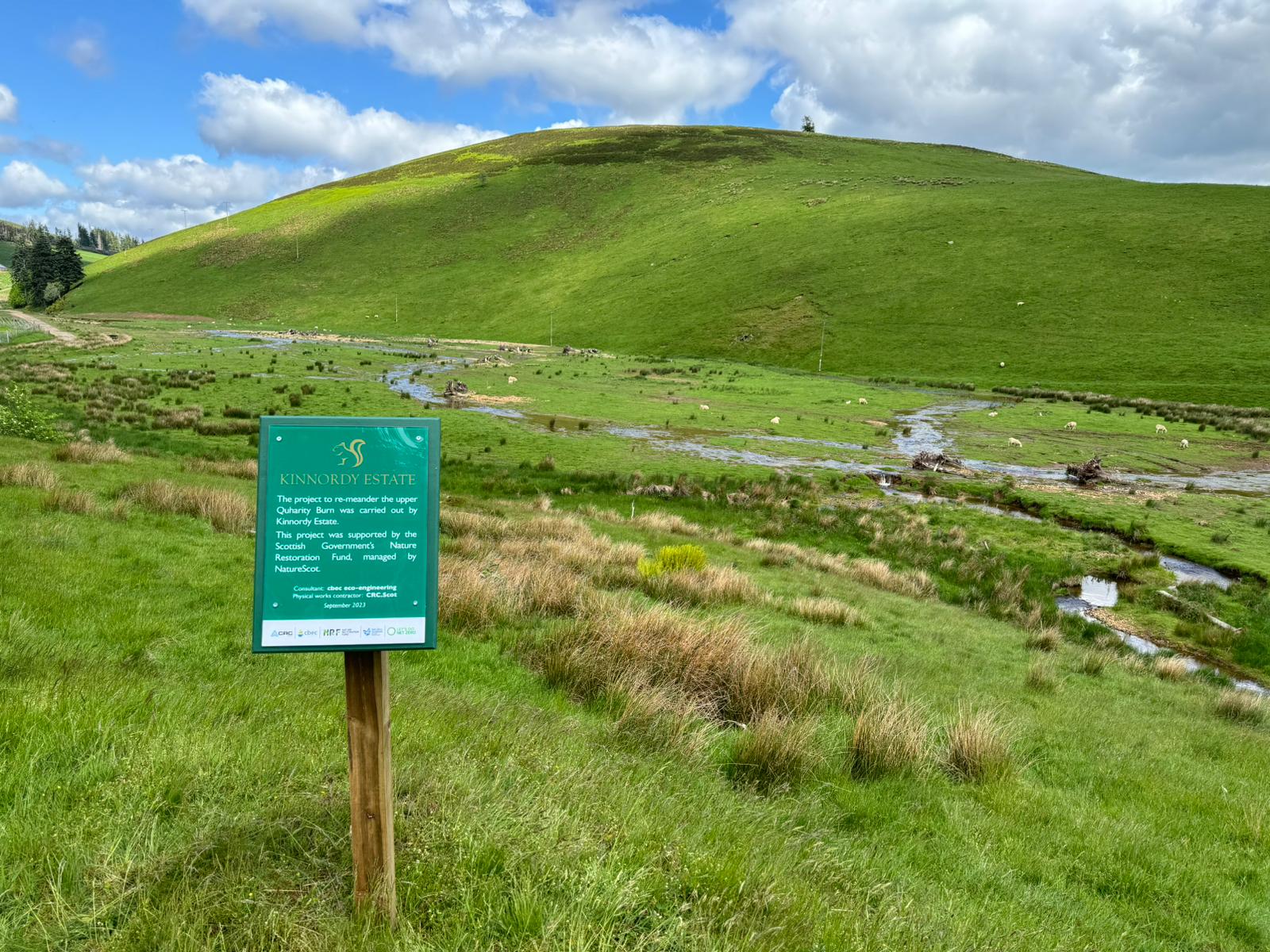 Scottish moorland with small hill or elevation in the background and in the foreground a printed sign identifying this as the area of the Quharity burn restoration project