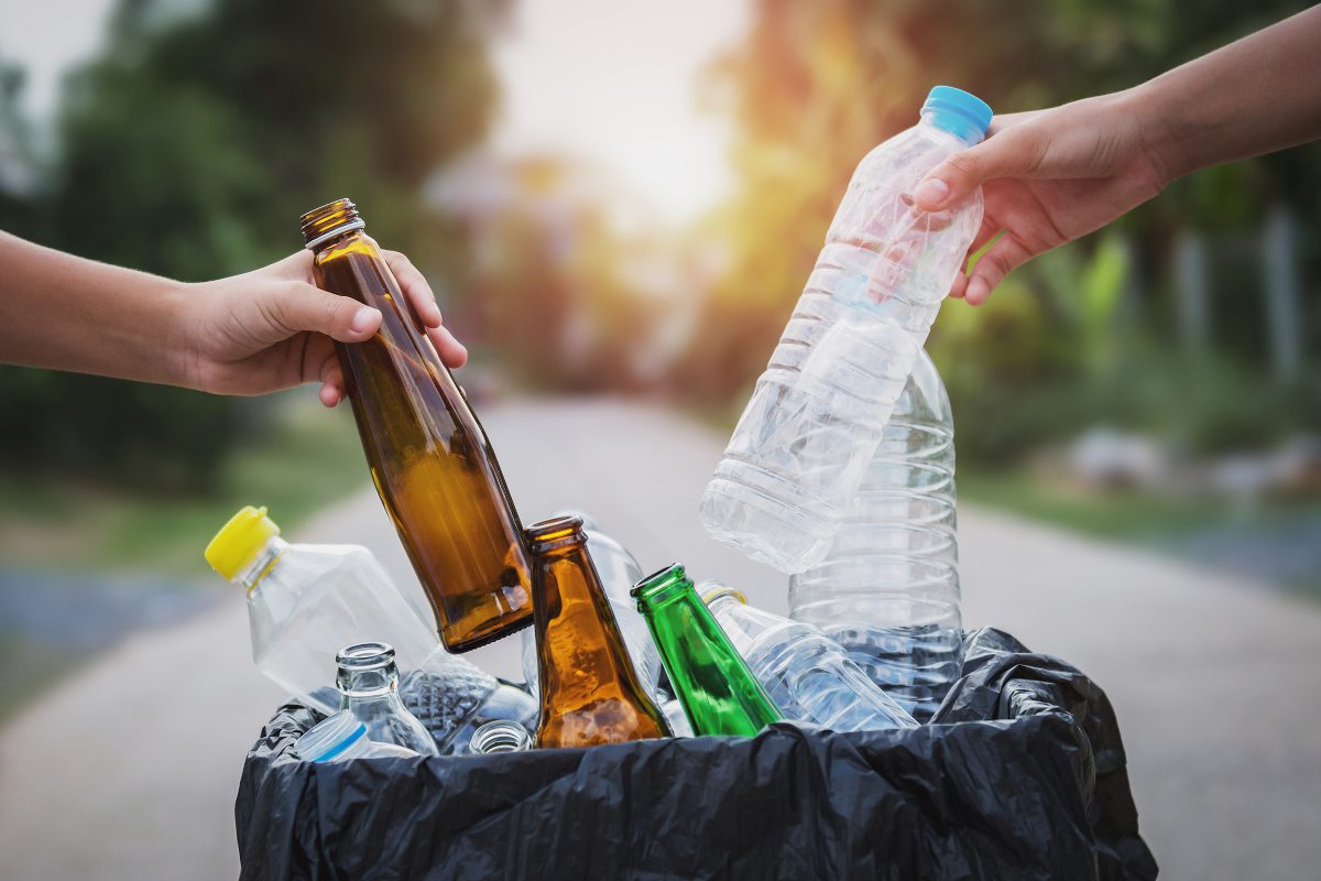 Hands place glass and plastic bottles into a recycling collection receptacle with background suggesting a household driveway location