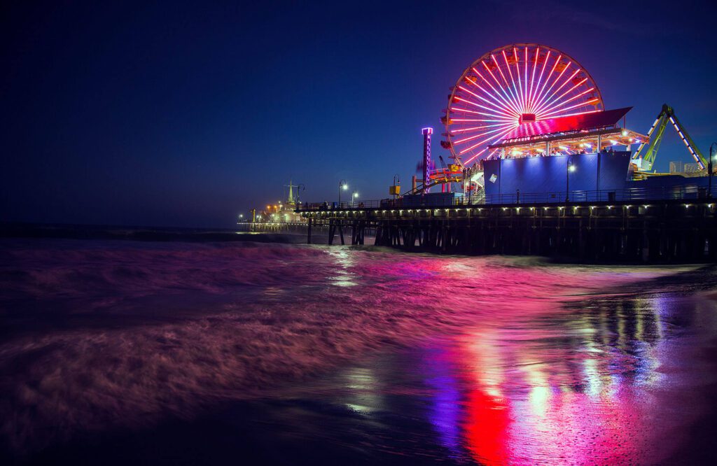 Nighttime beach scene with pier in the background atop which are amusements and illuminated big wheel