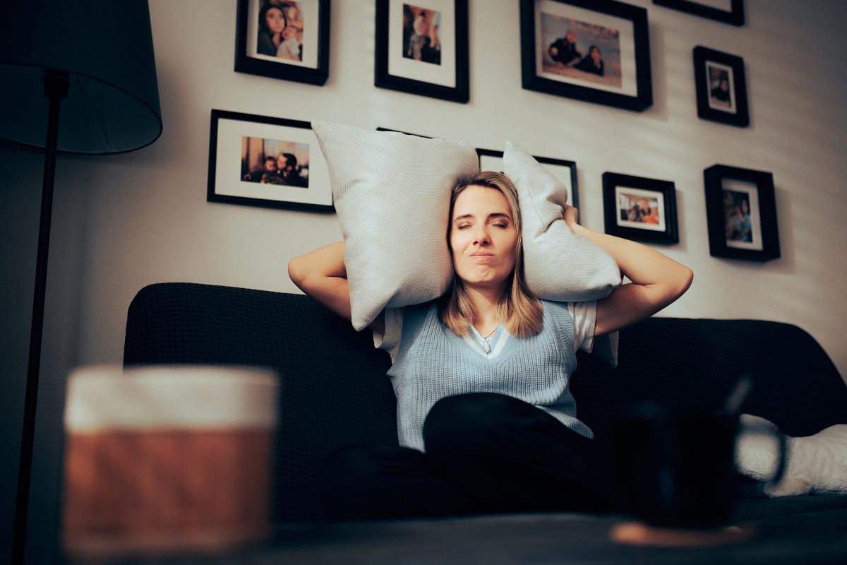 A woman on a sofa wrapping a pillow around her head to drown out noise