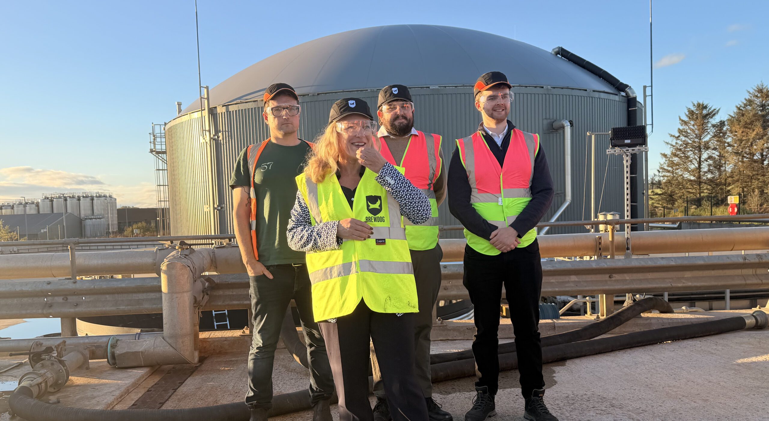 A group of people in hi-vis vests over normal clothing stand outdoors with a large digester dome in the background, a hundred metres or so behind them, with a blue sky overhead and trees and buildings also visible in the background