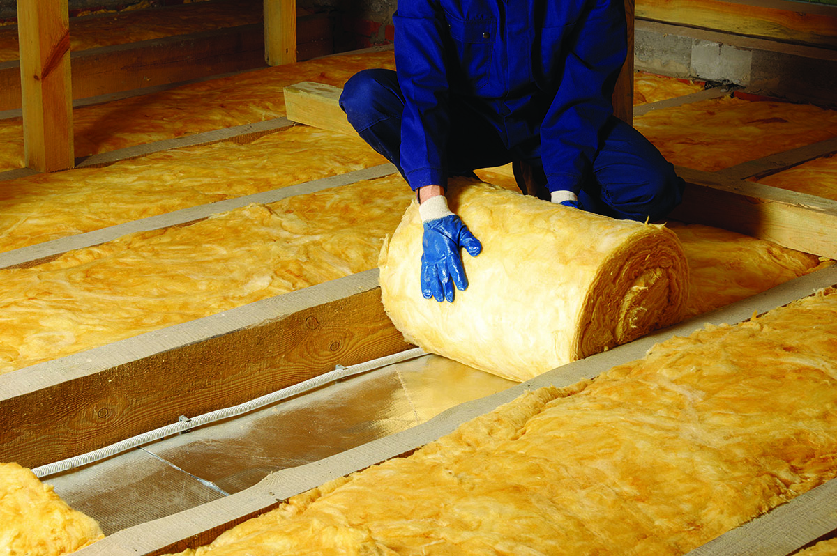 Orange fluffy cavity insulation being unrolled onto a flooring space by a blue-boiler-suited person whose squatting lower body and arms are visible