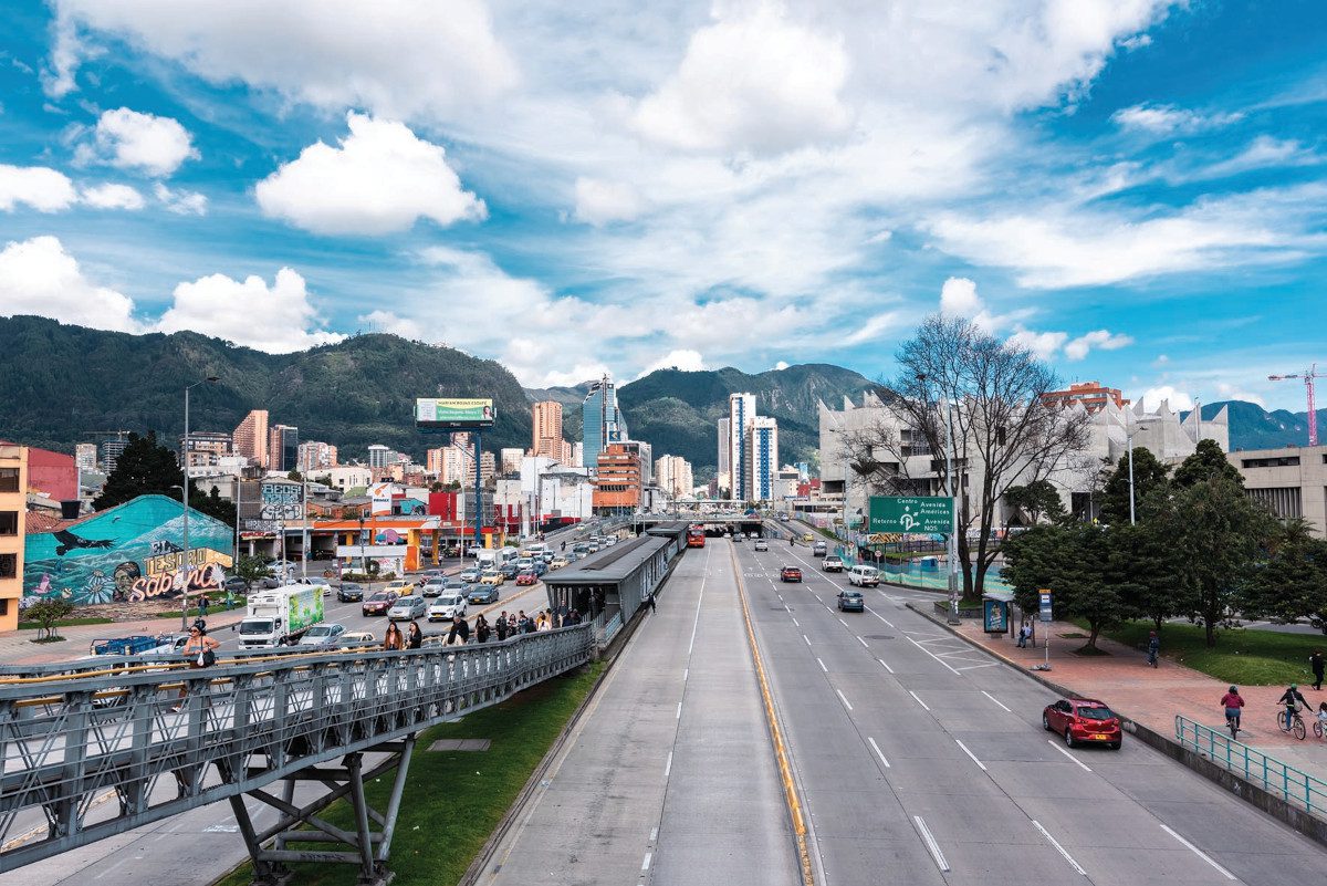View down a freeway which leads off in the distance towards a modest cityscape, with some tall buildings, all framed by dark- or green-coloured shapes of mountains and blue sky overhead with some cloud cover
