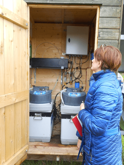 Woman in winter jacket seen in profile examining interior of wooden hut in which are visible cylindrical tanks, tubes and other equipment