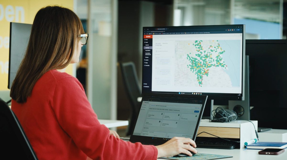 A woman in a red jumper, seen in profile, works at a desktop computer, the screen on which shows a dashboard with a map marked with coloured blobs