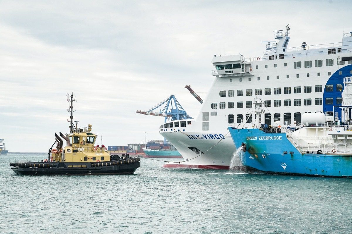 A passenger ferry with a green-coloured hull is floating in a location resembling a port or harbour, while nearby there is also a white tourism-related vessel and a smaller vessel, possibly a refuelling craft, which is between and in proximity to the two larger vessels