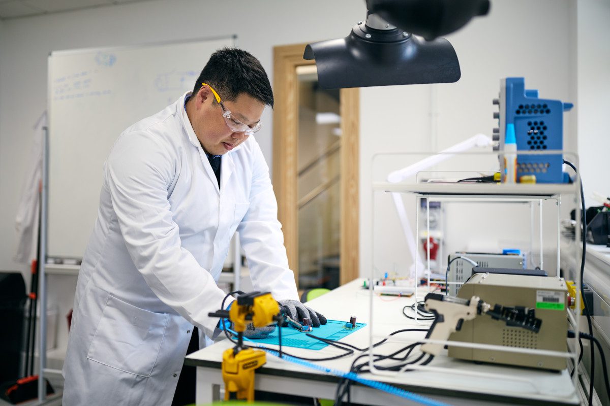 White-coated man in laboratory setting looking down at a benchtop upon which hi-tech equipment is laid out