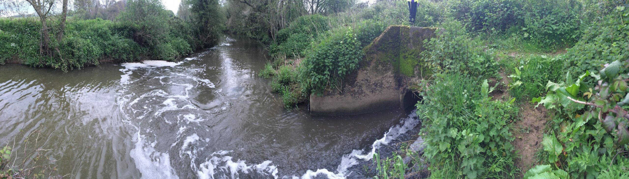 A portion of a river in a seemingly rural setting with a culvert or similar stone structure visible near the water's edge