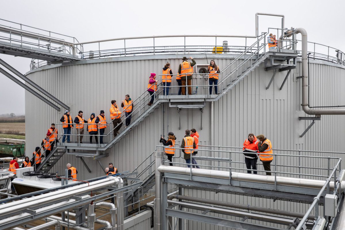 a large cylindrical biogas tank, the sides of which are lined with stairways, upon which are lined official delegates and visitors wearing hi-vis