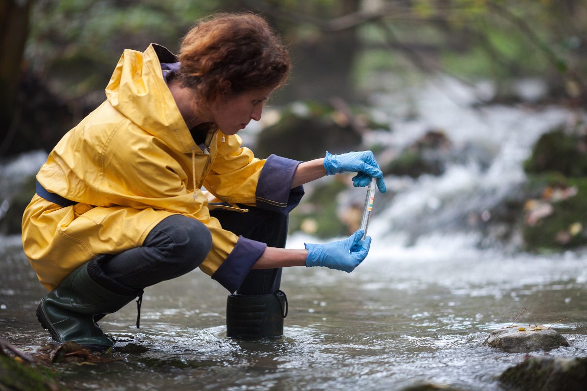 A woman - a scientist - in a low stance, collecting water samples from a river