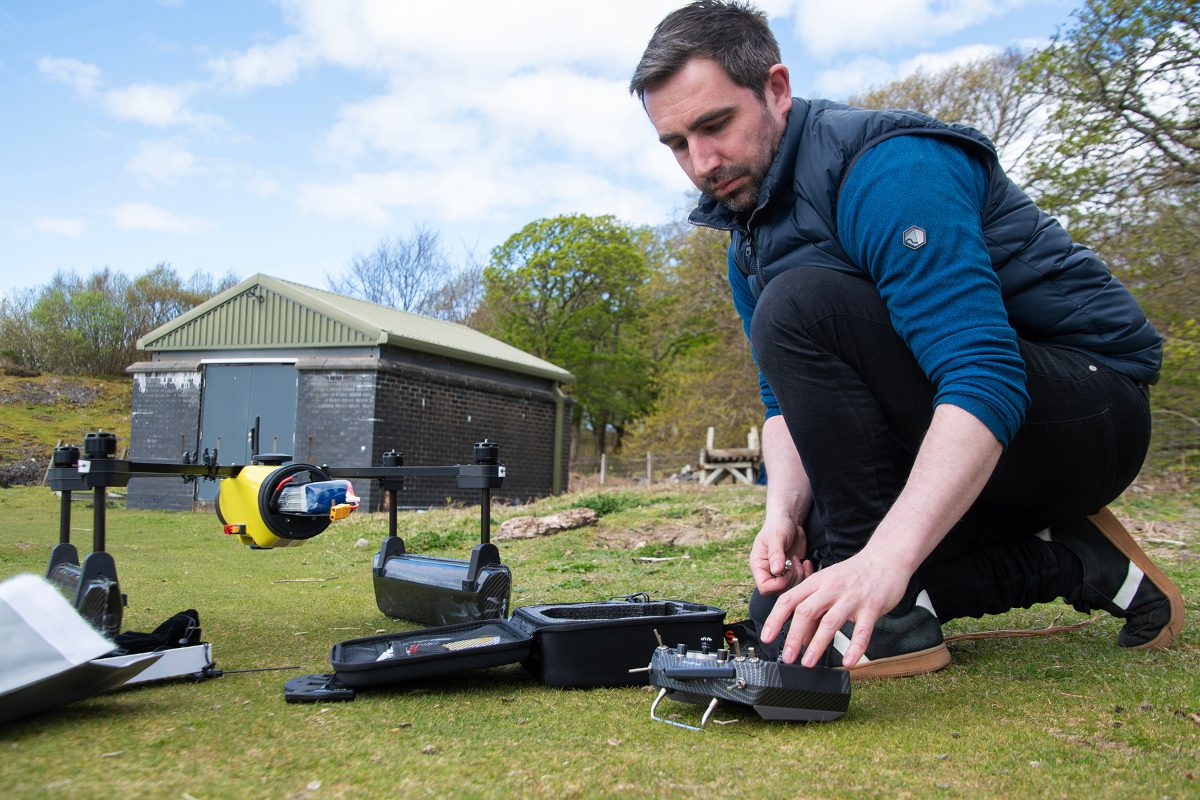 A man crouches down with one hand extended downwards to adjust or move a small object, an autonomous vehicle, all in an outdoor location