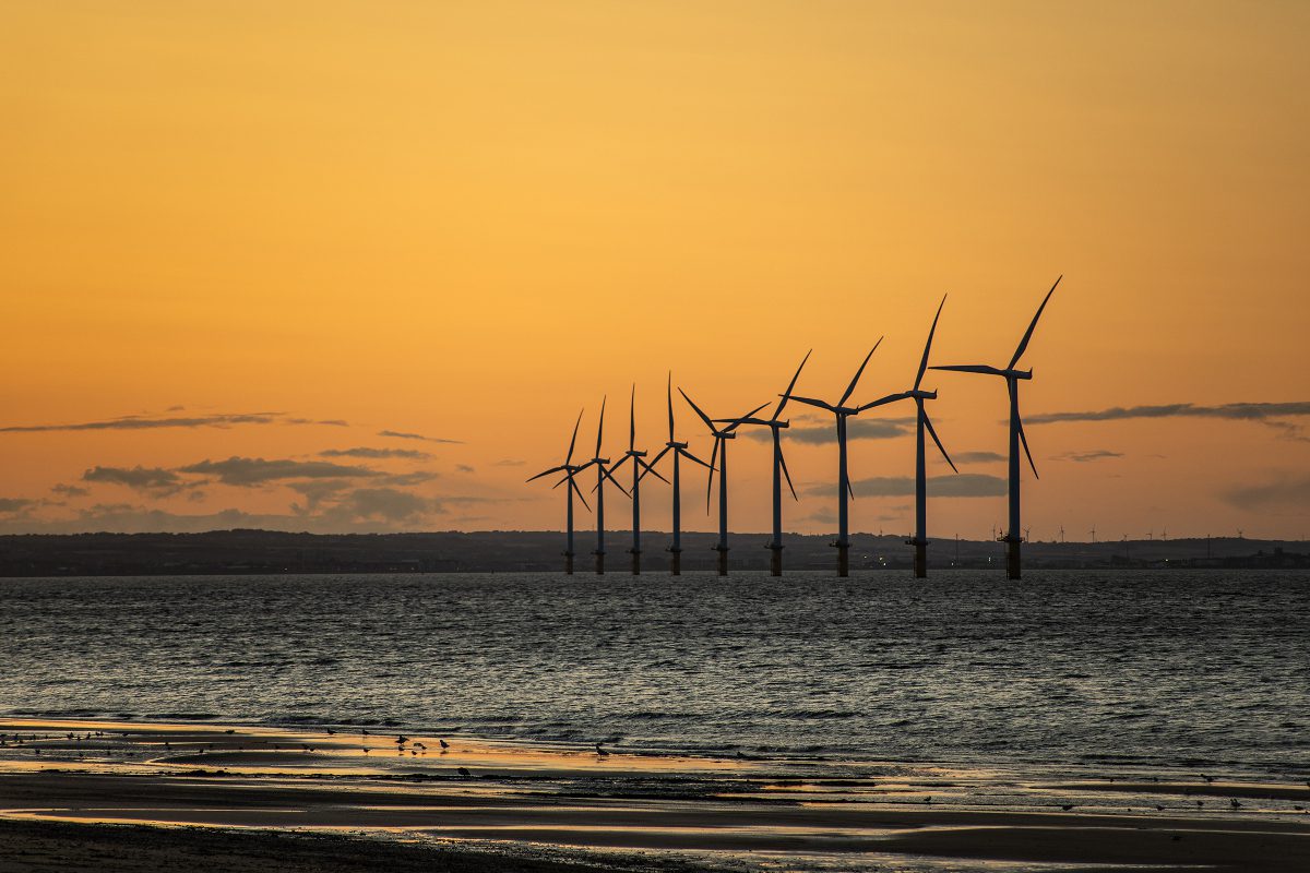 A row of offshore wind turbines in the distance, seen from the shoreline, visible as silhouettes against sunset