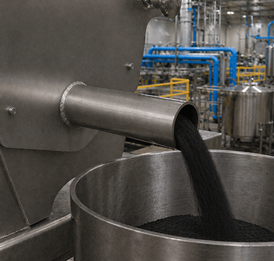 Close-up of an industrial stainless steel outlet pipe dispensing black granular material into a metal container, with processing equipment and blue piping visible in the background.