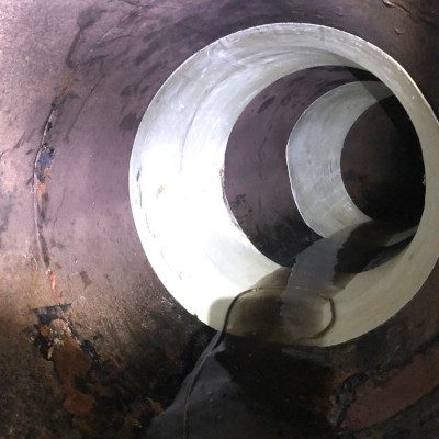 Close-up view inside a large corroded pipe showing a newly installed white liner segment, with water flowing along the bottom.