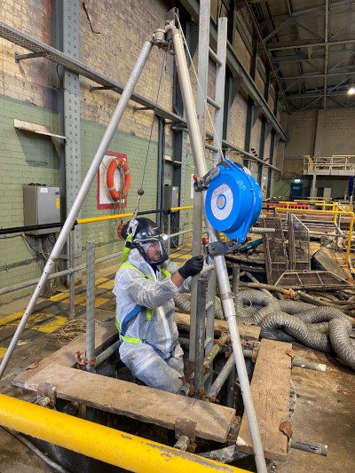 Operative in specialist waterproof gear including sealed facemask on metal ladder which is protruding from a space in the floor inside the interior of a building