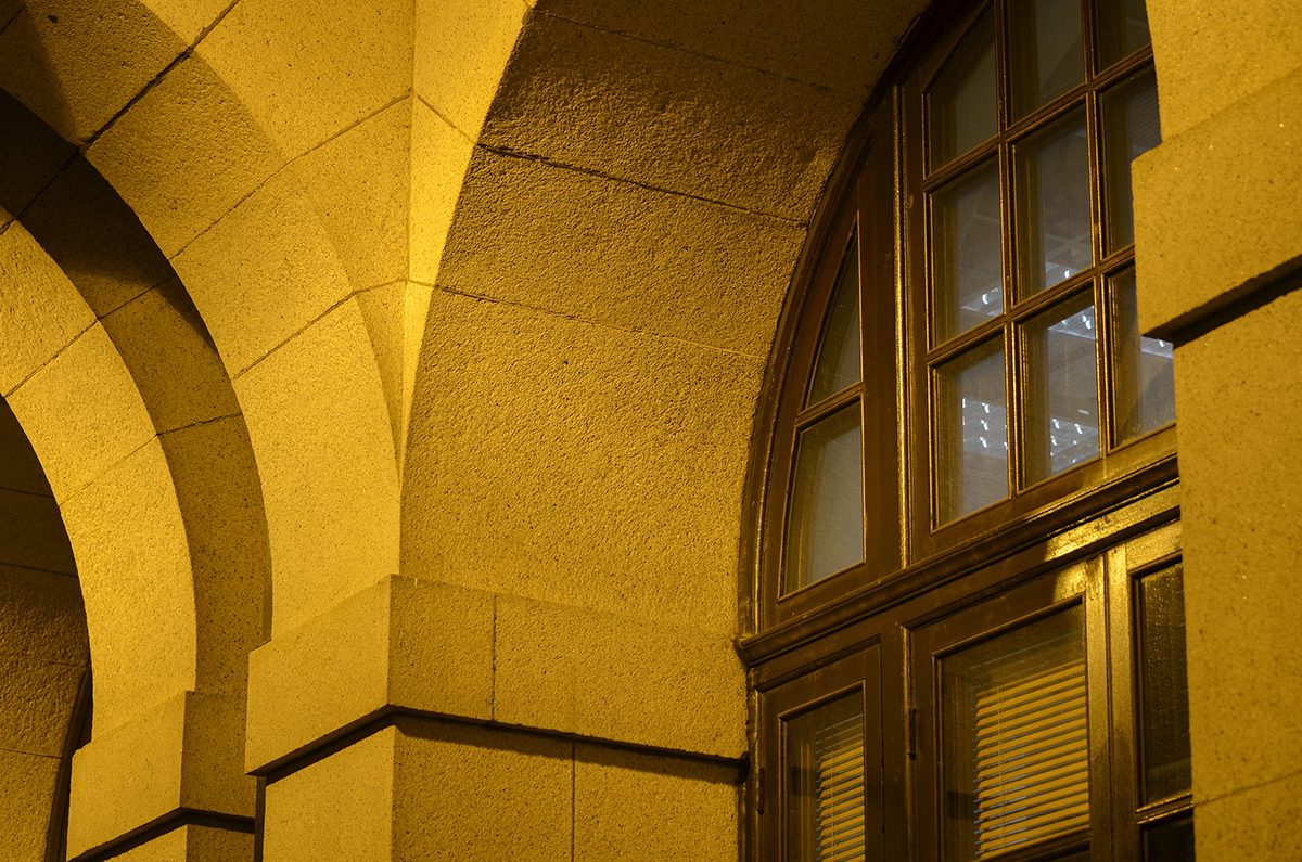 Close-up of a stone building façade with arched columns and a large wooden-framed window, illuminated by warm yellow lighting at night