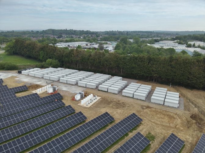 Aerial view of the Copwood VFB Energy Hub in East Sussex, showing rows of solar panels in the foreground alongside dozens of white vanadium flow battery units arranged in a grid beside a tree line.