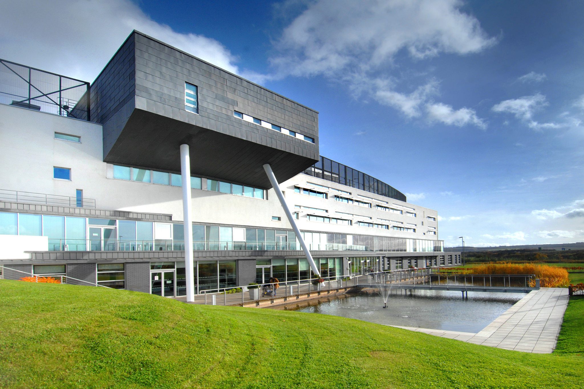 Modern university campus building with contemporary black and white façade, elevated angular structure supported by columns, glass-fronted walkways and landscaped grounds surrounding a water feature under a blue sky.