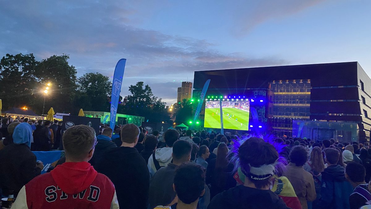 Football fans watch a game on a big screen in a nighttime urban location