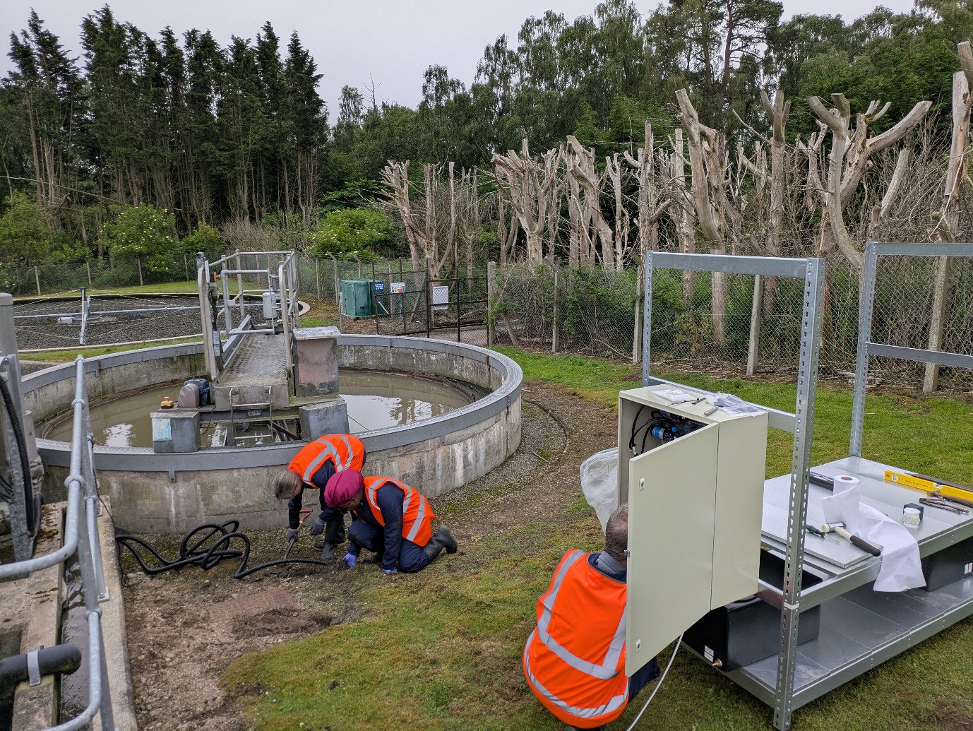 Researchers in high-visibility clothing installing monitoring and electrical equipment beside a circular wastewater treatment tank at a Scottish Water treatment works
