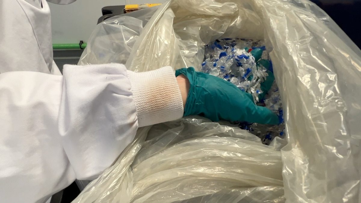 Gloved laboratory worker handling shredded plastic waste collected in a large bag.