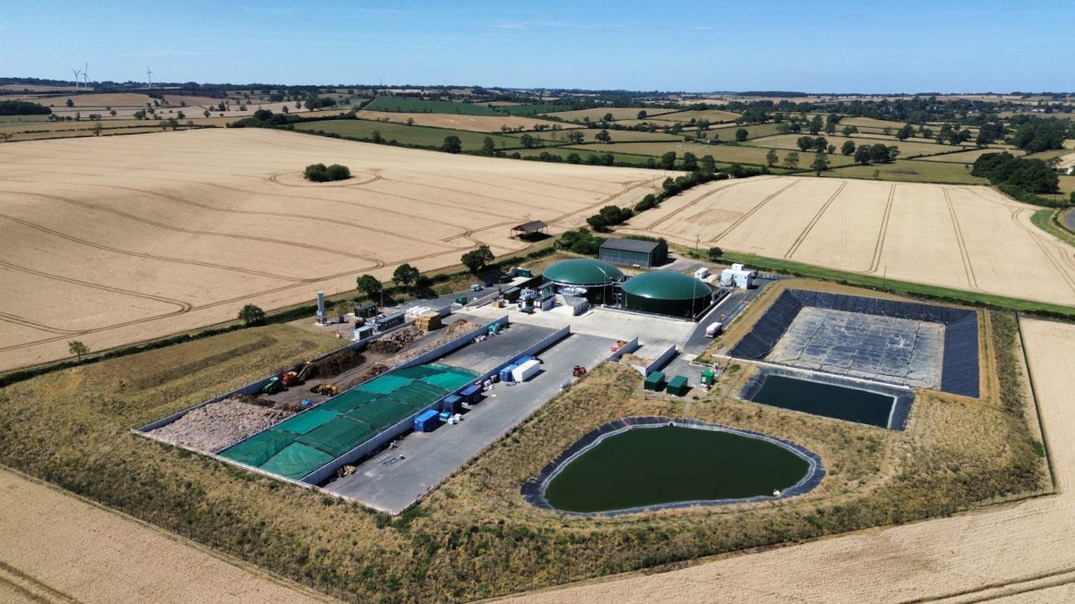 Aerial view of a biogas production facility surrounded by agricultural fields, showing two large green-domed tanks, covered storage bays, lined lagoons and associated plant equipment.
