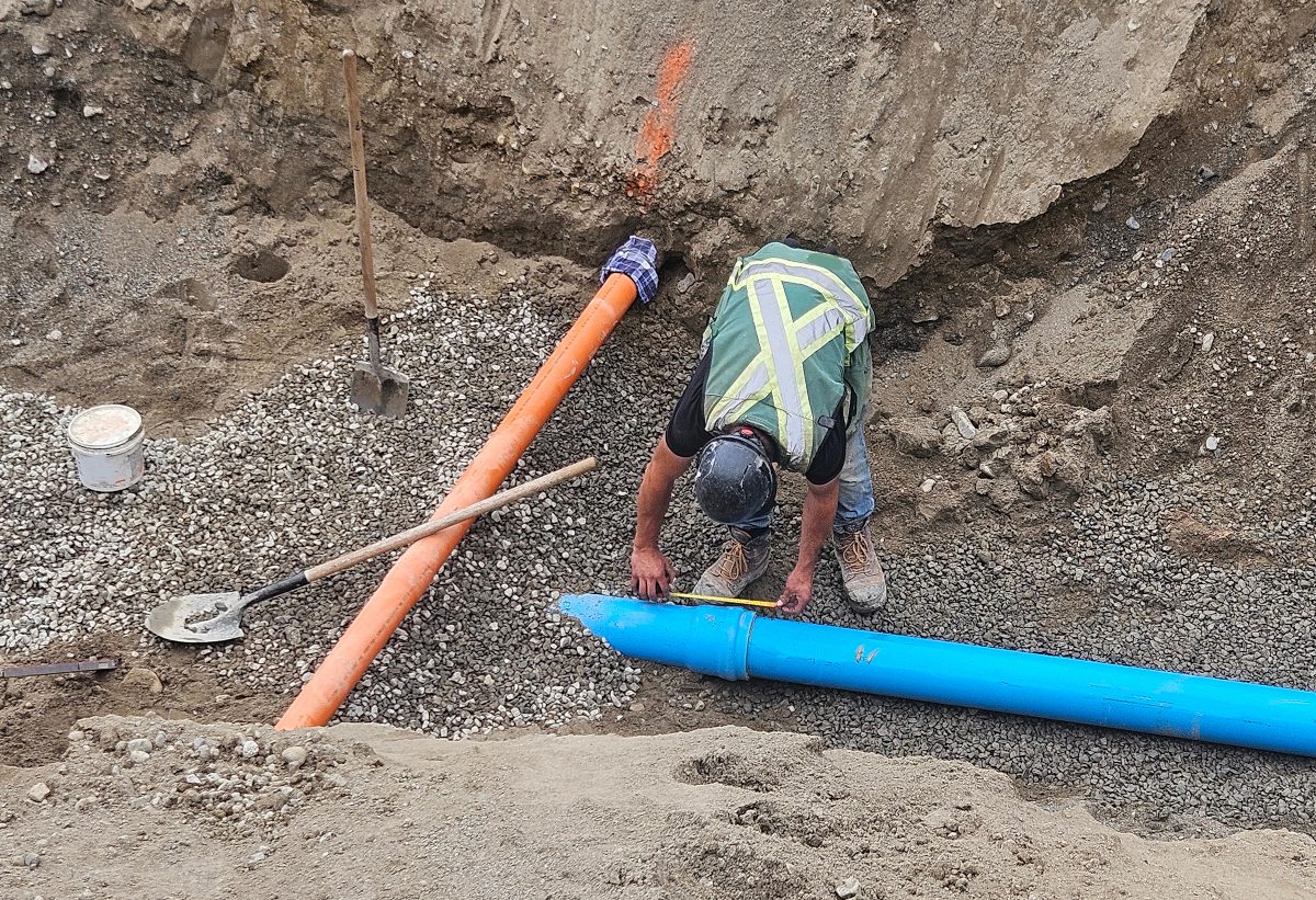 Construction worker wearing a hard hat and high-visibility vest positioning a blue pipe in an excavated trench alongside an orange pipe and gravel bedding.