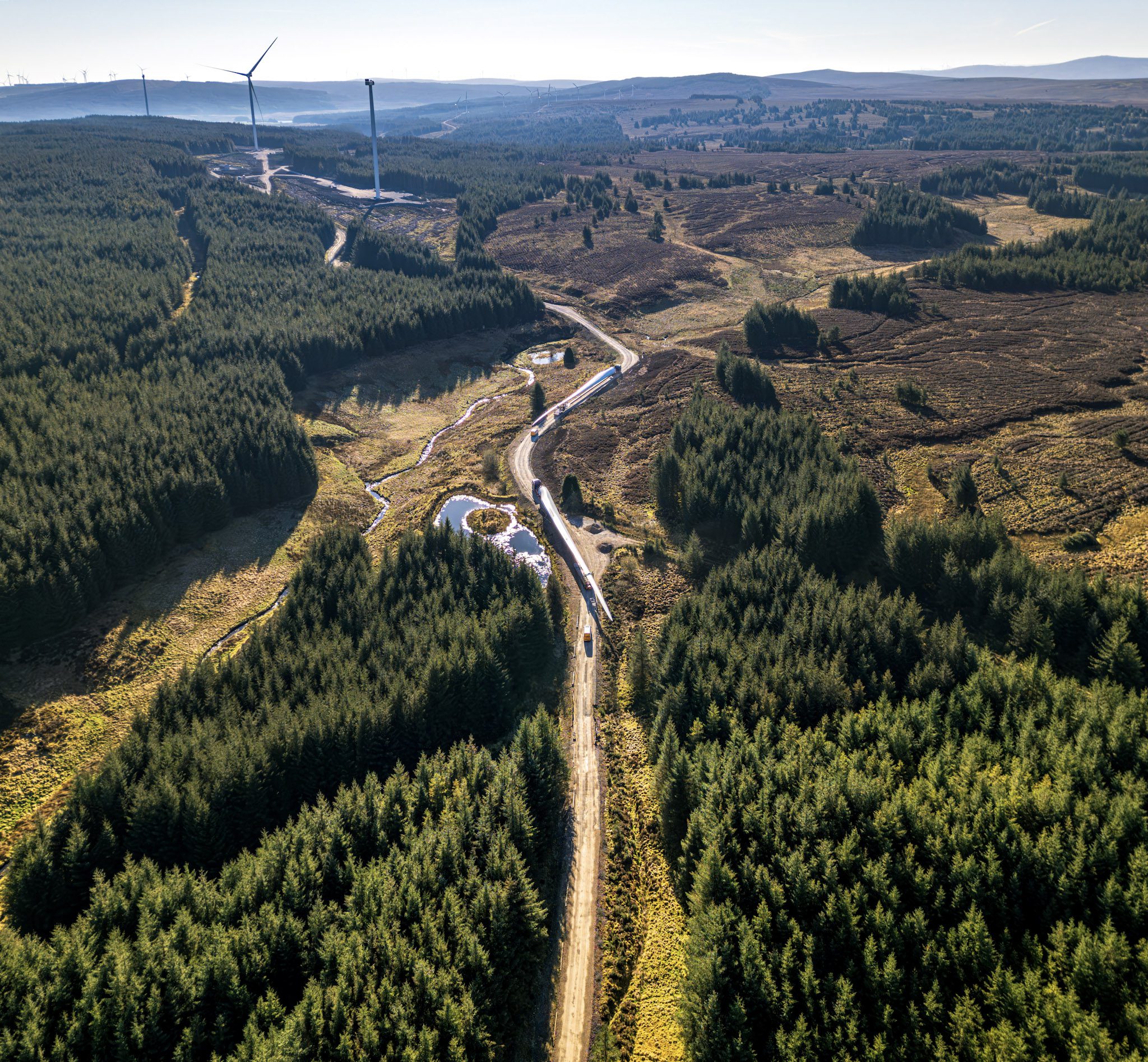 Aerial view of a convoy transporting long wind turbine blades along a winding access road through forested countryside, with wind turbines visible on the surrounding hills