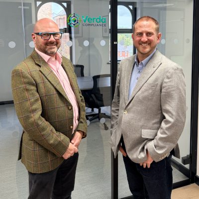 Two men standing in an office in front of a glass meeting room displaying the Verda Compliance logo